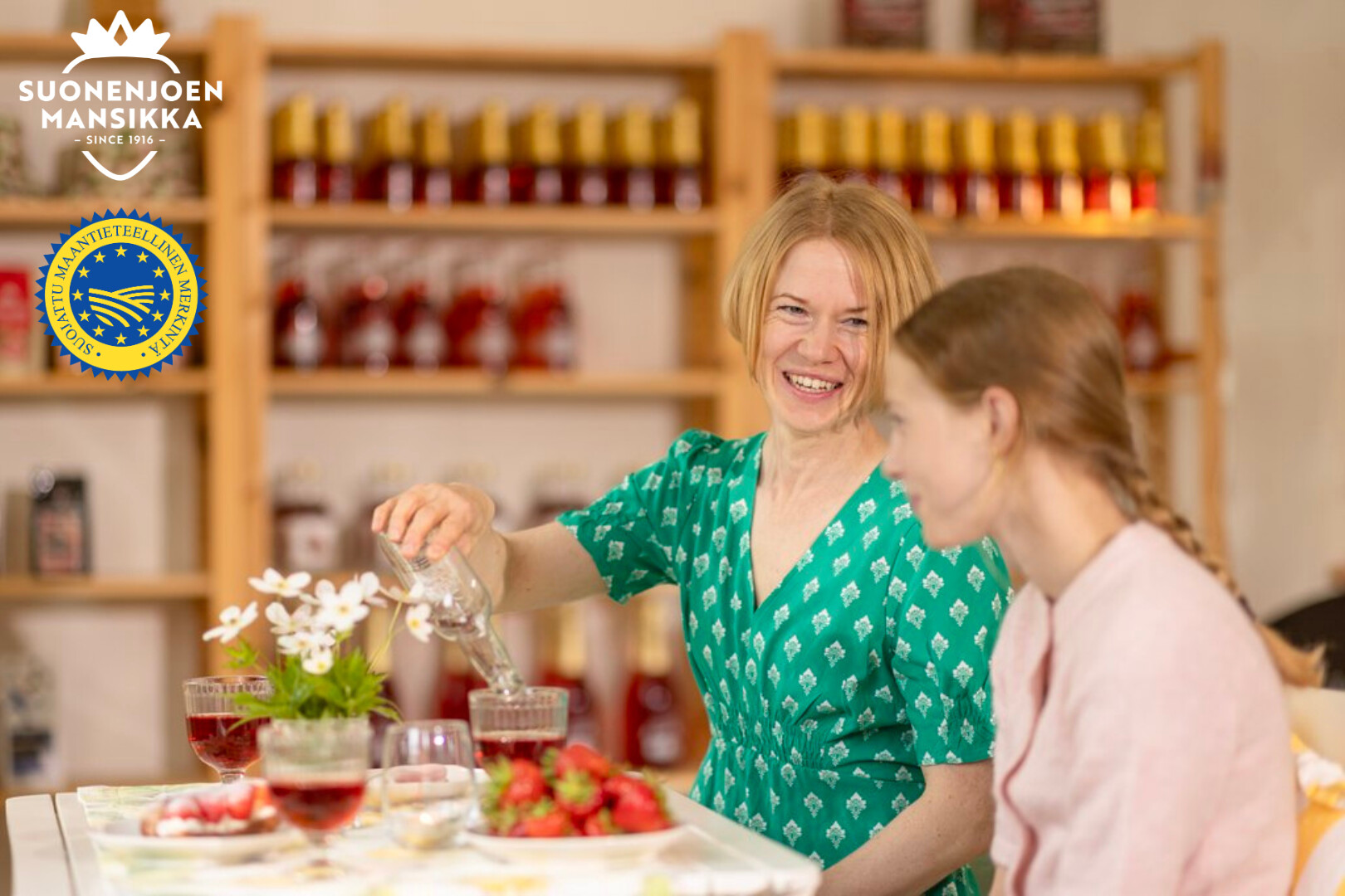 Kaksi naista istuu pöydässä, jossa on mansikoita, juomia ja kukkia. Toinen vihreäpukuinen nainen kaataa vettä hymyillen. Taustalla on hyllyjä, joissa on pulloja. Logot näkyvät vasemmassa yläkulmassa.