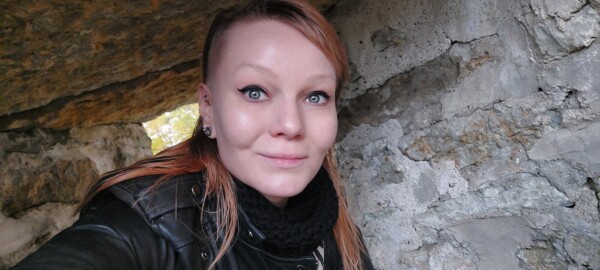 Vaaleaihoinen, punatukkainen ja sinisilmäinen nainen hymyilee kameralle seisoessaan ulkona karhean kiviseinän vieressä. Hänellä on yllään musta huivi ja musta nahkatakki.