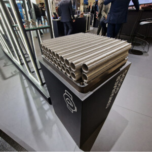 Pino metalliputkia, joissa on uurrettuja pintoja ja jotka ovat esillä mustalla jalustalla sisätiloissa järjestettävässä näyttelyssä, ja taustalla näkyy ihmisiä ja moderneja kojuja.