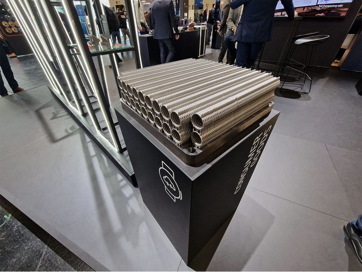 Pino metalliputkia, joissa on uurrettuja pintoja ja jotka ovat esillä mustalla jalustalla sisätiloissa järjestettävässä näyttelyssä, ja taustalla näkyy ihmisiä ja moderneja kojuja.