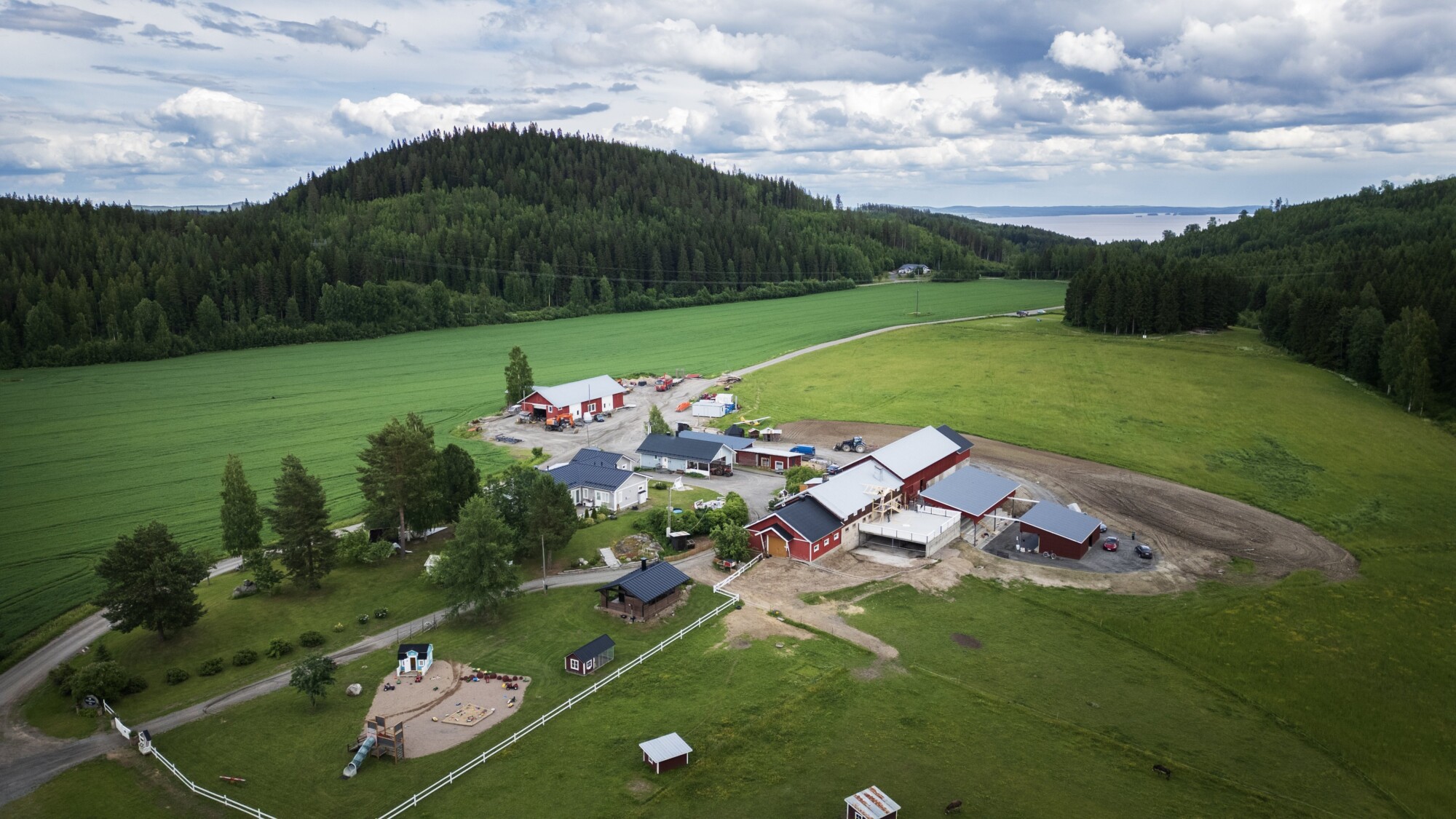 Ilmakuva maaseudun maatilasta, jossa on punaisia ja valkoisia latoja, vihreiden peltojen, puiden ja kukkuloiden ympäröimänä pilvisen taivaan alla. Rakennusten lähellä näkyy leikkikenttä ja aidattuja alueita.