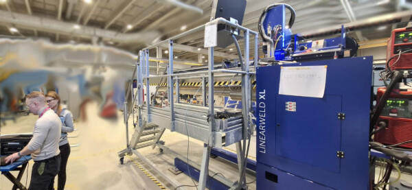 Kaksi ihmistä seisoo suuren teollisuuskoneen vieressä, jossa lukee LINEARWELD XL, tilavassa, hyvin valaistussa tehdasympäristössä. Koneessa on siniset ohjauspaneelit, ja metallikaiteet ympäröivät aluetta.
