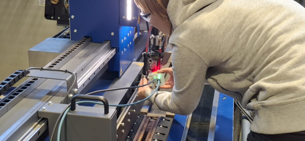 Harmaaseen huppariin pukeutunut henkilö säätää anturia tai laitetta sinisessä teollisuuskoneessa, johon on kytketty kaapeleita ja jonka laitteessa palaa punainen valo.