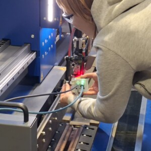 Harmaaseen huppariin pukeutunut henkilö säätää anturia tai laitetta sinisessä teollisuuskoneessa, johon on kytketty kaapeleita ja jonka laitteessa palaa punainen valo.