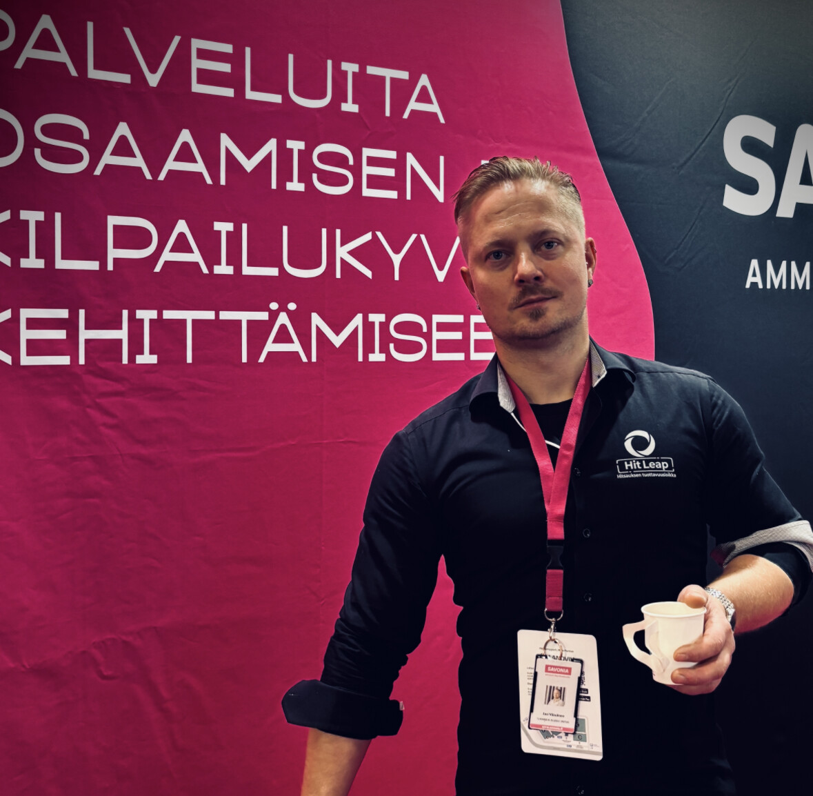 Mustaan paitaan ja nimimerkkiin pukeutunut mies seisoo kuppi kädessään näytön edessä, jossa on iso suomenkielinen teksti vaaleanpunaisella ja mustalla pohjalla.