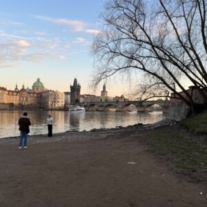 Kaksi ihmistä seisoo joen rannalla Prahassa, taustalla historiallinen Kaarlensilta ja kaupungin rakennuksia, ja oikealla puolella on suuri lehdetön puu. Taivas on osittain pilvinen auringonlaskun aikaan.