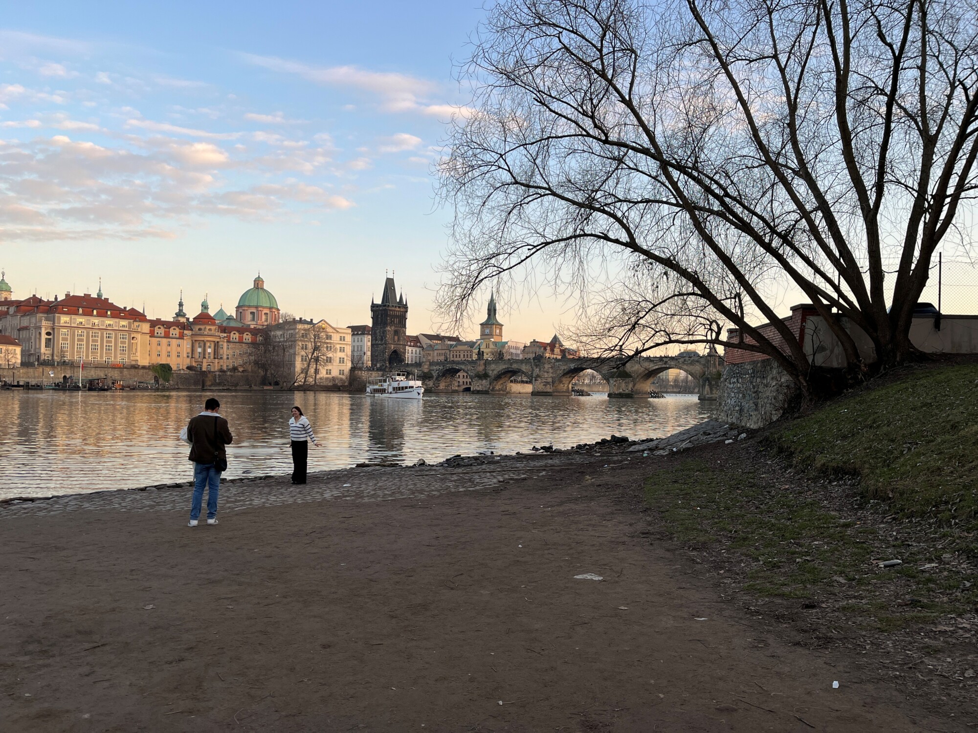 Kaksi ihmistä seisoo joen rannalla Prahassa, taustalla historiallinen Kaarlensilta ja kaupungin rakennuksia, ja oikealla puolella on suuri lehdetön puu. Taivas on osittain pilvinen auringonlaskun aikaan.