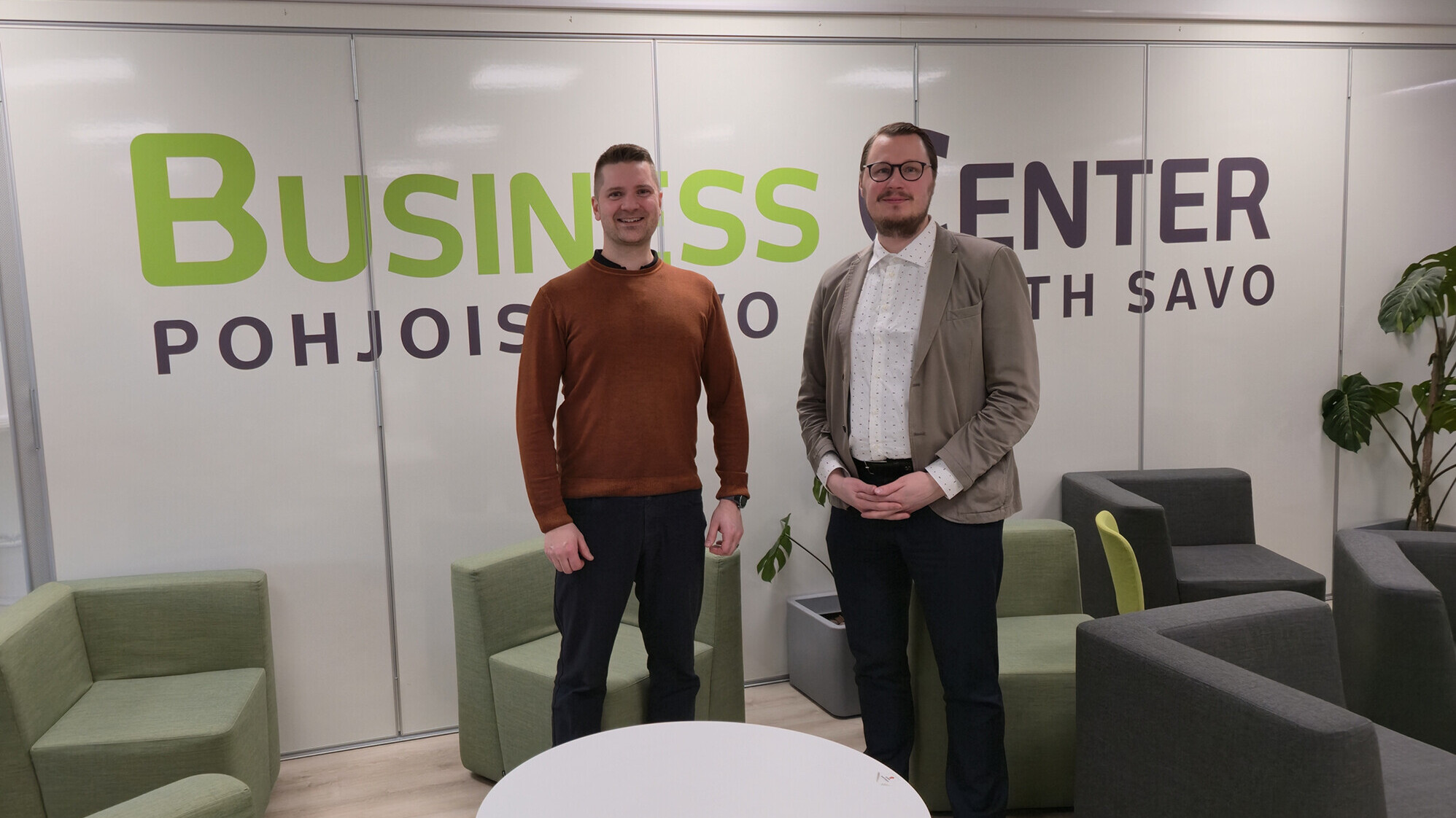 Kaksi miestä seisoo kameraan päin modernissa toimistohuoneessa, jossa on vihreitä ja harmaita tuoleja. Heidän takanaan on seinä, jossa lukee Business Center Pohjois-Savo Etelä-Savo.