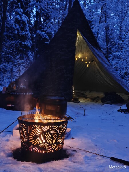 Kuvioitu nuotiopaikka palaa lumessa lähellä telttaa, joka on valaistu valonjohdoilla lumisessa metsässä iltahämärässä. Ruukut ovat nuotion päällä ja lähellä, ja ne luovat viihtyisän talvisen leirintäalueen.