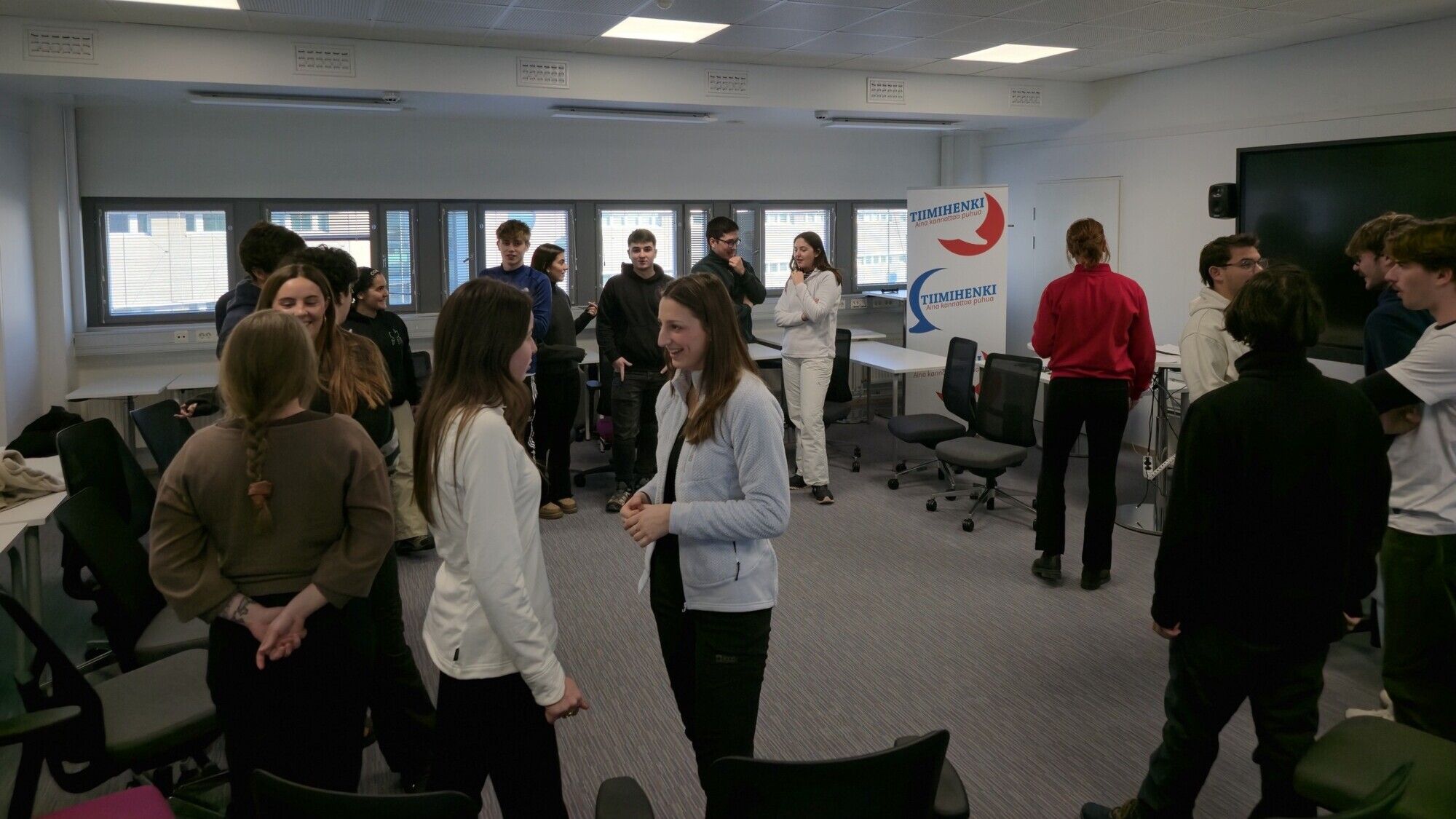 Joukko nuoria seisoo ja keskustelee pienissä ryhmissä valoisassa, modernissa luokkahuoneessa, jossa on tuoleja ja pöytiä. Taustalla näkyy kyltti, jossa on sinisiä ja punaisia nuolia ja sana Thomasius.