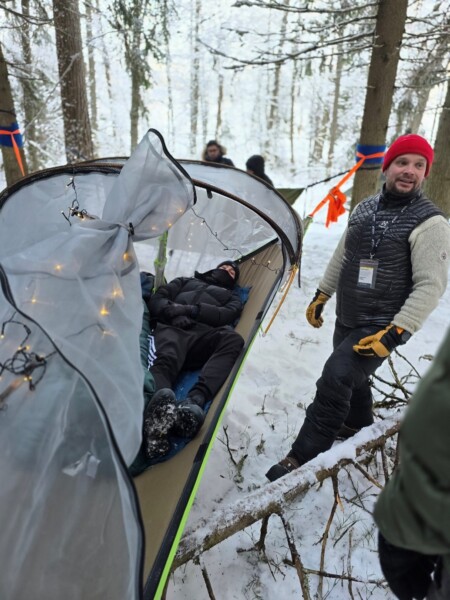 Henkilö makaa riipputeltassa tai riippumatossa, jossa on verkkopeite ja valonjohto, lumen peittämien puiden ympäröimänä. Toinen henkilö talvivaatteissa ja punaisessa pipossa seisoo lähellä ja hymyilee.