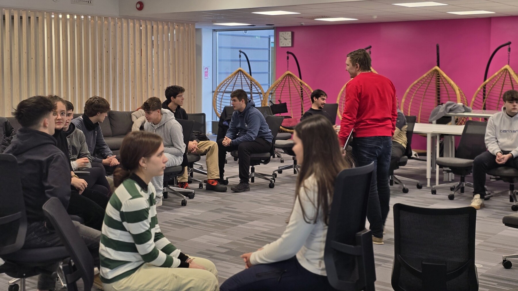 Ryhmä ihmisiä istuu pareittain toimistotuolilla keskustelemassa modernissa huoneessa, jossa on vaaleanpunaiset seinät, roikkuvat tuolit ja suuret ikkunat. Yksi henkilö seisoo, kun muut istuvat ja keskustelevat.