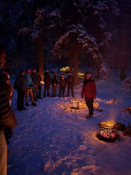 Joukko talvivaatteisiin pukeutuneita ihmisiä seisoo yöllisessä lumisessa metsässä ja on kokoontunut nuotiopaikkojen ympärille, jotka heittävät lämpimän hehkun lumeen ja puihin.
