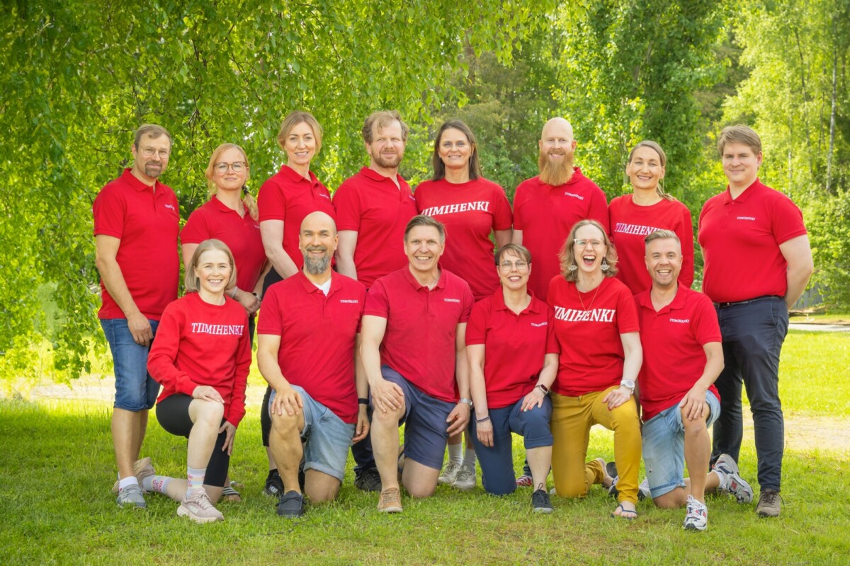Kolmentoista aikuisen ryhmä poseeraa ulkona vihreällä nurmikolla, kaikilla on yllään samanlaiset punaiset paidat, joissa lukee "TIIMIHENKI", ja shortsit tai farkut, ja he hymyilevät lehtivihreiden puiden edessä aurinkoisena päivänä.