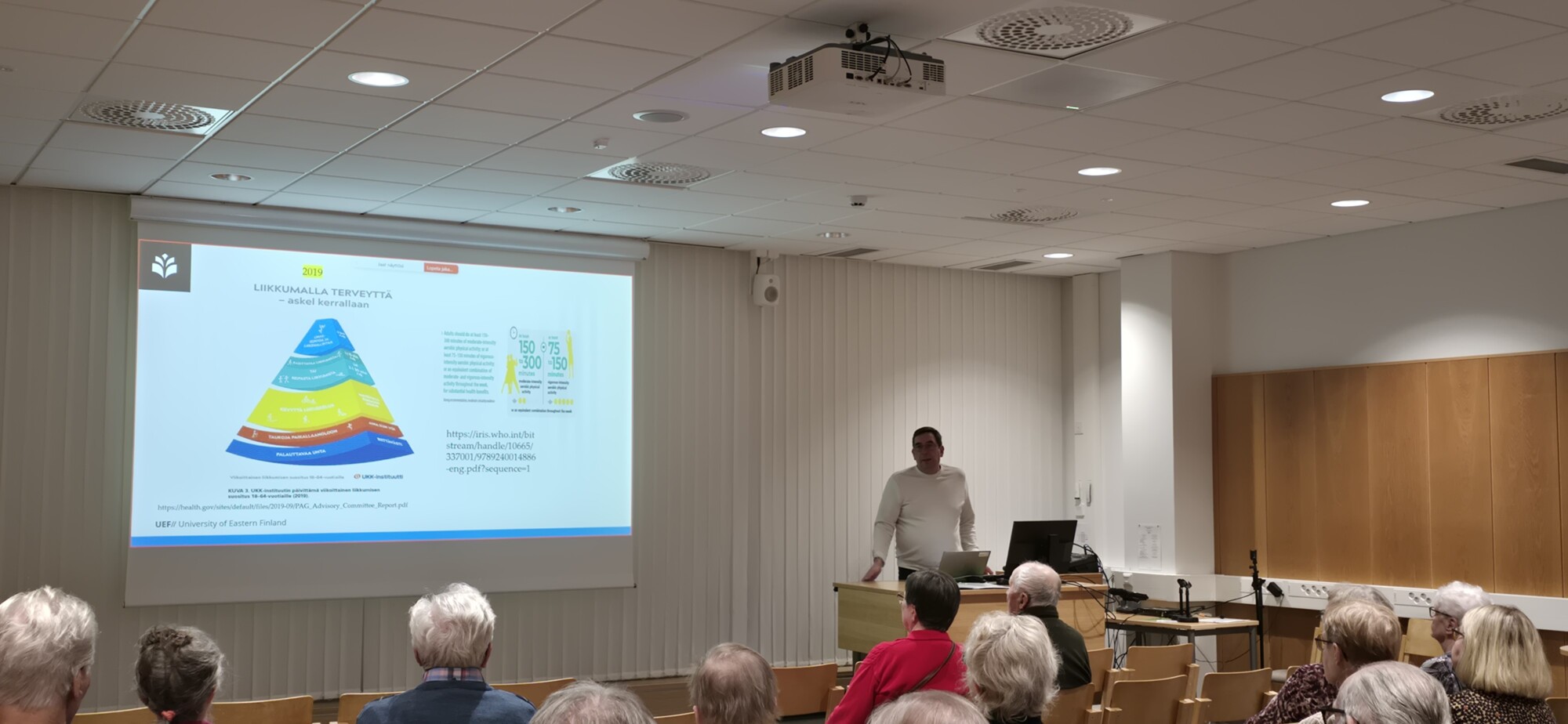 Mies seisoo korokkeella pitämässä esitelmää luokkahuoneessa olevalle yleisölle. Pyramidikaavio ja teksti heijastetaan huoneen etuosassa olevalle näytölle. Yleisö istuu paikallaan ja on tarkkaavainen.