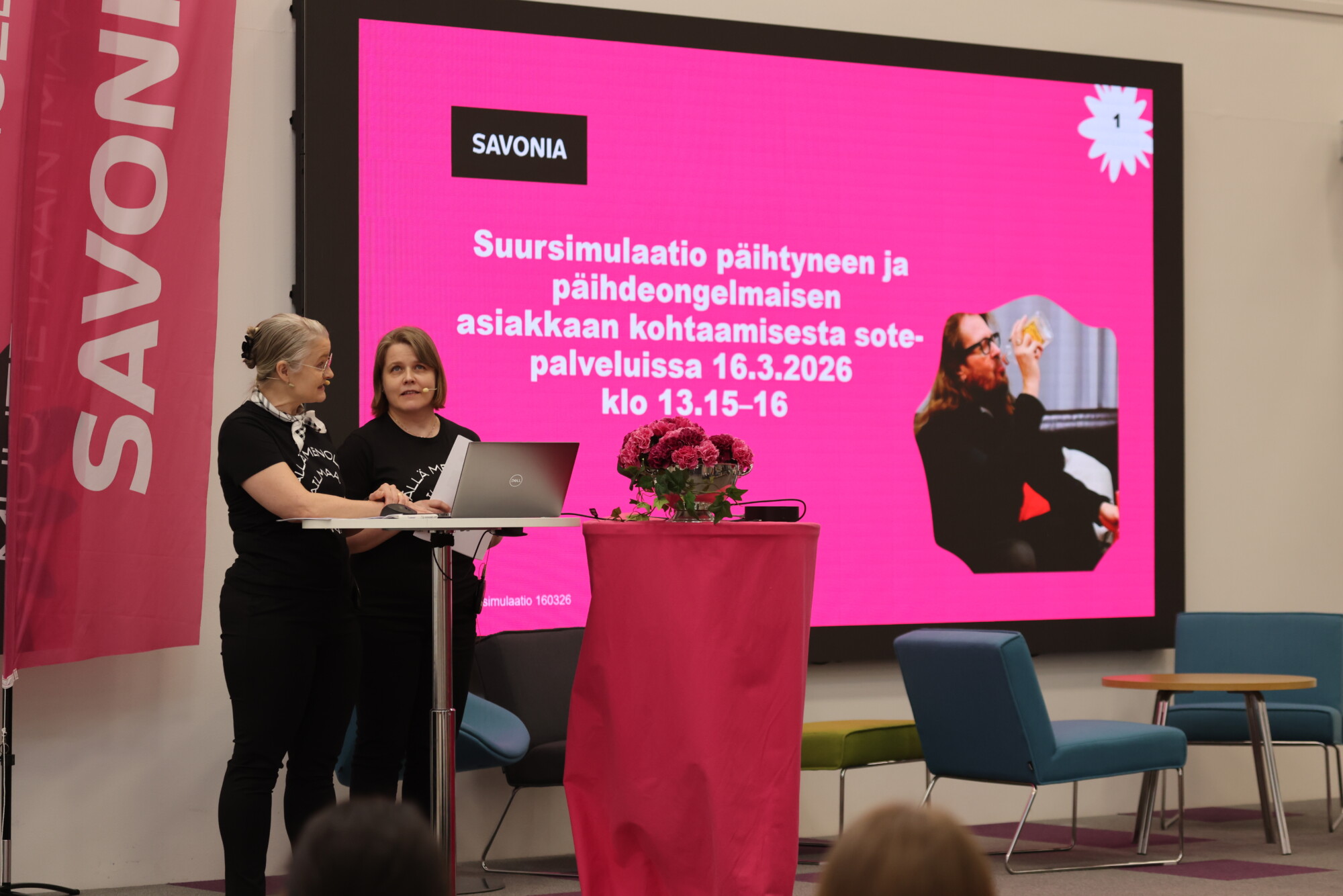 Kaksi naista seisoo korokkeen ääressä esittelemässä tapahtumaa. Heidän takanaan on suuri vaaleanpunainen näyttö, jossa on valkoinen suomenkielinen teksti, Savonia-logo ja kuva viittomakieltä käyttävästä henkilöstä. Näkyvissä on vaaleanpunainen lippu ja kukkakimppu.
