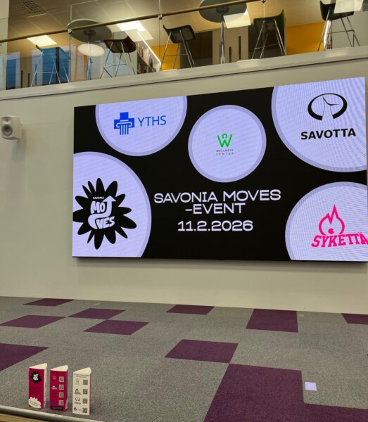 Suurella digitaalisella näytöllä näkyy "SAVONIA MOVES - TAPAHTUMA 11.2.2026" ja eri organisaatioiden logoja matolla päällystetyn lava-alueen yläpuolella; edessä lattialla on kolme pientä seisovaa kylttiä.