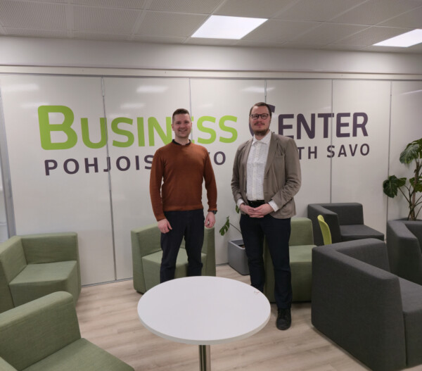 Kaksi miestä seisoo modernissa toimiston oleskelutilassa, jossa on vihreitä ja harmaita tuoleja. Heidän takanaan seinällä on teksti Business Center Pohjois-Savo Pohjois-Savo.
