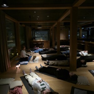 Ryhmä ihmisiä makaa matoilla hämärässä huoneessa ja osallistuu rentoutus- tai meditaatioistuntoon. Rauhallista ilmapiiriä lisäävät kynttilät, tyynyt ja muutama lattialle asetettu kulho. Suurista ikkunoista avautuu öinen kaupunkinäkymä.