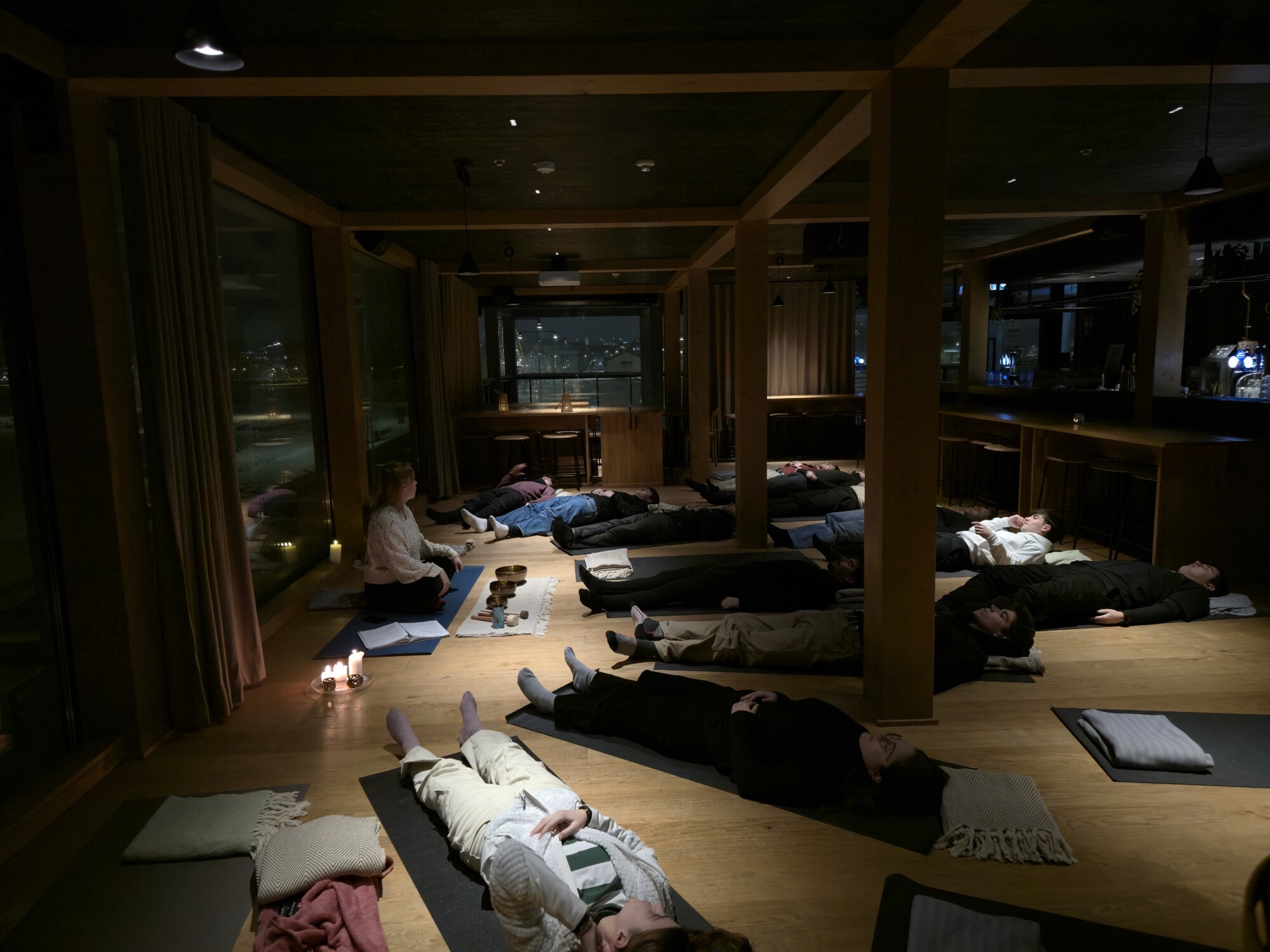 Ryhmä ihmisiä makaa matoilla hämärässä huoneessa ja osallistuu rentoutus- tai meditaatioistuntoon. Rauhallista ilmapiiriä lisäävät kynttilät, tyynyt ja muutama lattialle asetettu kulho. Suurista ikkunoista avautuu öinen kaupunkinäkymä.