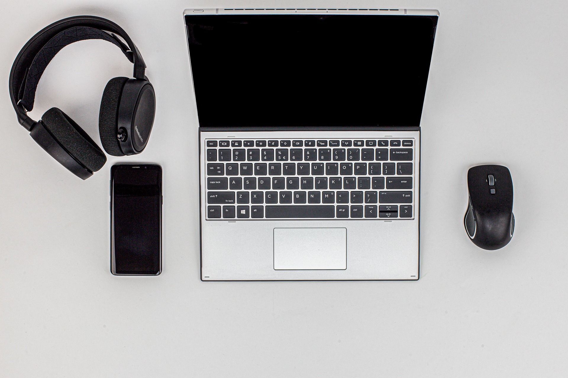 Hopeinen kannettava tietokone, jossa on musta näyttö, on keskellä valkoista pintaa, ja sitä reunustavat mustat langattomat kuulokkeet ja älypuhelin vasemmalla ja musta langaton hiiri oikealla.