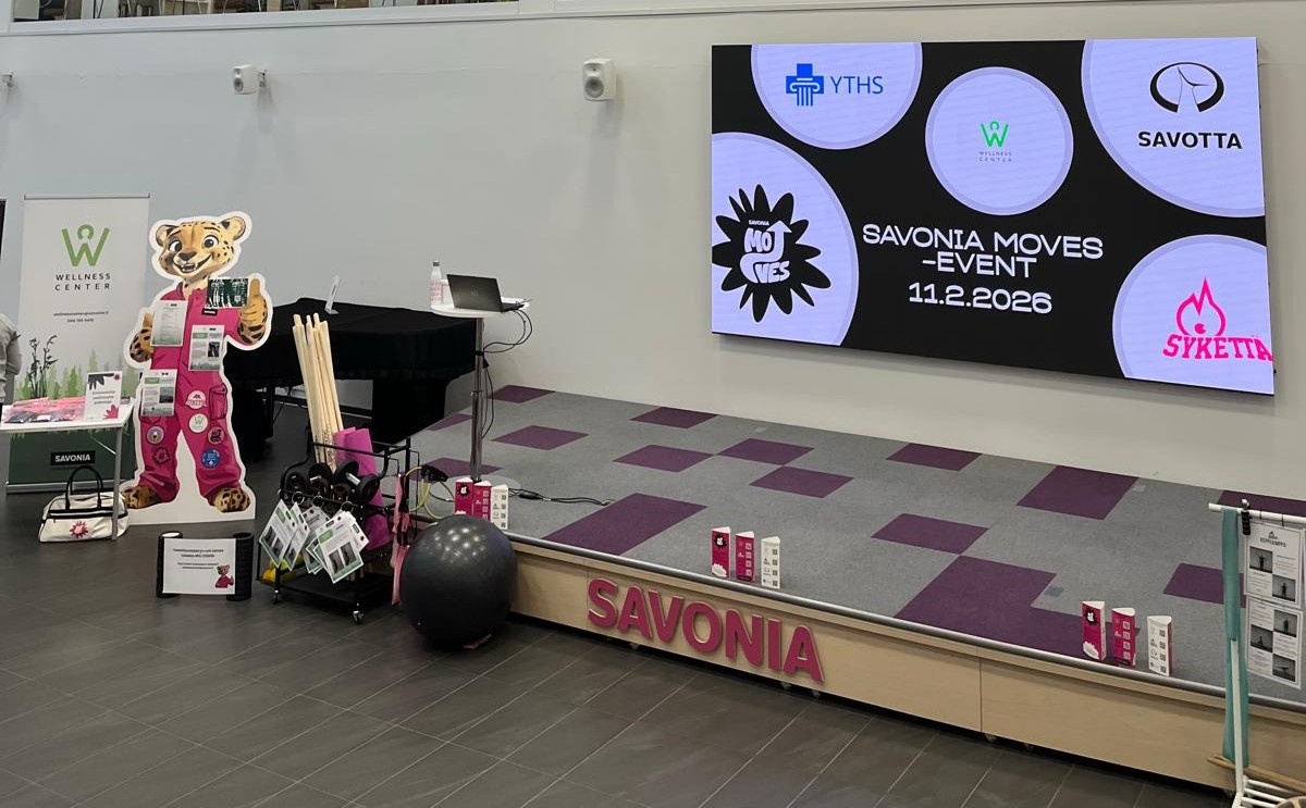 Sisätiloissa oleva tapahtumalava, jossa on suuri näyttö, jossa näkyy SAVONIA MOVES - TAPAHTUMA 11.2.2026. Lavan läheisyyteen on sijoitettu tiikerimaskotti, kuntoiluvälineitä, mainostuotteita ja bannereita. Sana SAVONIA on lavan etupuolella.