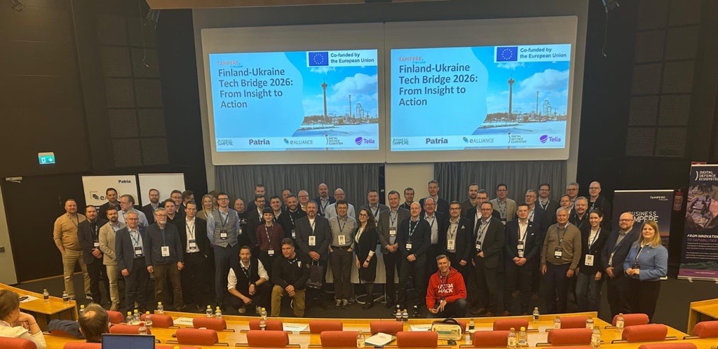 Suuri joukko ihmisiä poseeraa valokuvaa varten auditoriossa. Heidän takanaan kahdella näytöllä näkyy otsikko Finland-Ukraine Tech Bridge 2026: From Insight to Action sekä useita logoja ja kaupunkikuvia.
