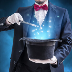 Taikuri, jolla on puku ja punainen rusetti, pitää taikasauvaa mustan silinterihatun päällä, ja hatusta nousee esiin sinisiä, kimaltelevia taikatehosteita tummaa, savuista taustaa vasten.