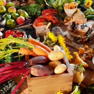 Värikäs valikoima tuoreita vihanneksia, hedelmiä ja gourmet-ruokia puisella pöydällä, mukaan lukien porkkanat, perunat, omenat, marjat, punajuuret, sienet, merenelävät, leivonnaiset ja keltaiset kukat koristeeksi.