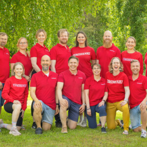 Kolmentoista aikuisen ryhmä, jolla on samanlaiset punaiset paidat, poseeraa ulkona nurmikolla, taustalla vihreitä puita. Jotkut seisovat, toiset polvistuvat, ja kaikki hymyilevät kameralle.