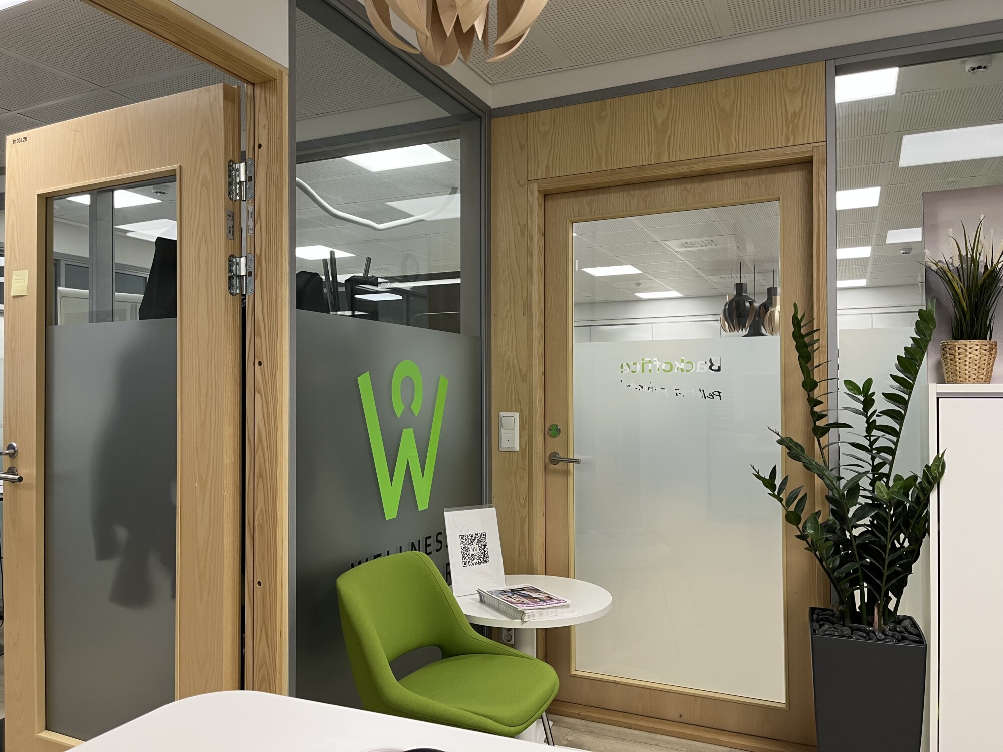 Moderni toimistotila, jossa on puuovet, huurrettu lasipaneeli, jossa on vihreä W-logo, vihreä tuoli pienen valkoisen pöydän vieressä, ruukkukasveja ja kattovalaisimet, jotka heijastuvat lasiin.
