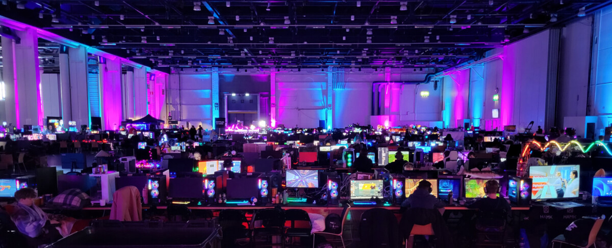 Suuri, hämärästi valaistu sali, joka on täynnä rivejä hehkuvia tietokonemonitoreja ja pelaavia ihmisiä, jotka ovat valaistu eloisilla sinisillä ja violeteilla valoilla ja luovat vilkkaan pelitapahtuman tunnelman.