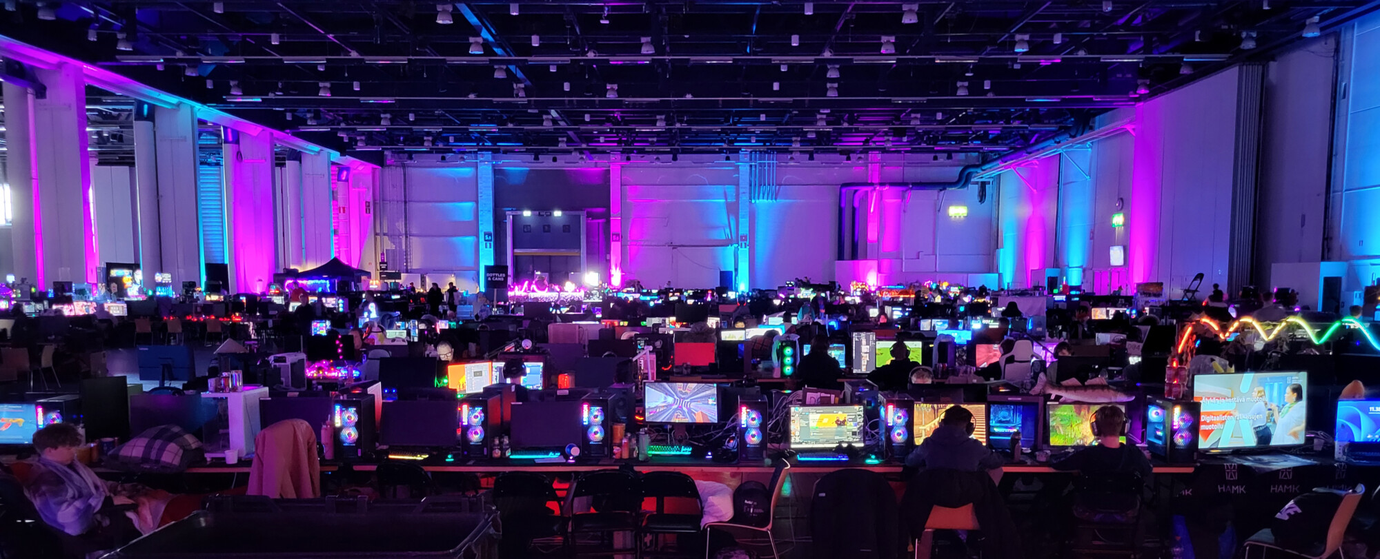 Suuri, hämärästi valaistu sali, joka on täynnä rivejä hehkuvia tietokonemonitoreja ja pelaavia ihmisiä, jotka ovat valaistu eloisilla sinisillä ja violeteilla valoilla ja luovat vilkkaan pelitapahtuman tunnelman.