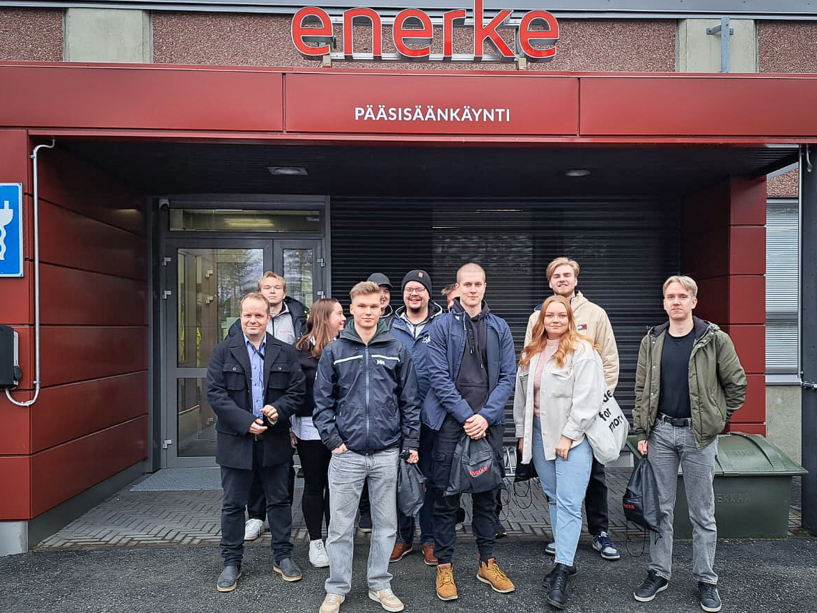 Yhdeksän hengen ryhmä seisoo punaisen rakennuksen sisäänkäynnin edessä, jonka oven yläpuolella on kyltti enerke ja Pääsisäänkäynti. Useimmilla on takit ja he näyttävät poseeraavan ryhmäkuvaa varten.