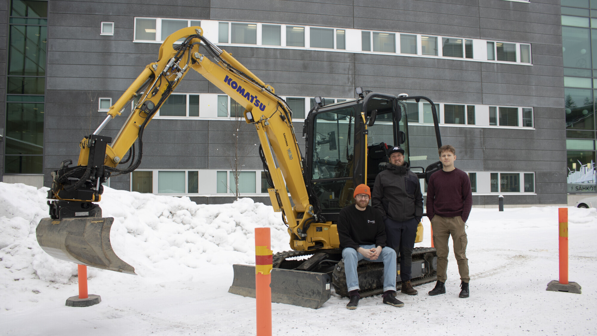 Kolme ihmistä seisoo pienen keltaisen Komatsu-kaivinkoneen edessä lumisella maalla, taustalla moderni rakennus ja lumikasoja. Etualalla näkyy kaksi oranssia pylvästä.