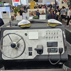 Lähikuva veneen kojelaudasta, jossa on ohjauspyörä, ohjauspaneelit, radio ja kaksi kapteeninhattua, ihmisten ympäröimänä kiireisessä sisätiloissa järjestettävässä näyttelyssä tai messutapahtumassa.