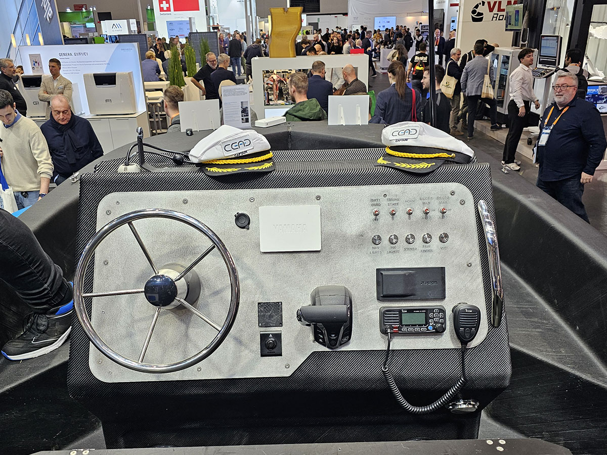 Lähikuva veneen kojelaudasta, jossa on ohjauspyörä, ohjauspaneelit, radio ja kaksi kapteeninhattua, ihmisten ympäröimänä kiireisessä sisätiloissa järjestettävässä näyttelyssä tai messutapahtumassa.
