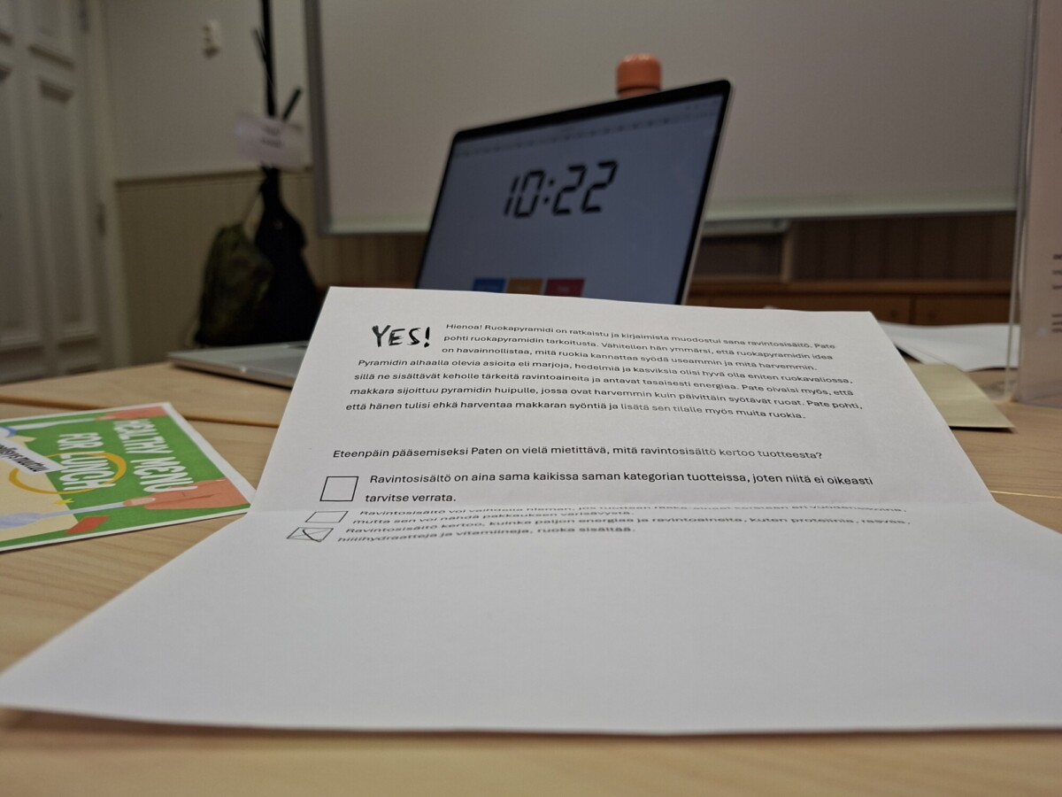 Lähikuva paperiarkista, jossa on suomenkielistä tekstiä kirjoituspöydällä, taustalla kannettava tietokone, jossa näkyy kello 10:22 ja muita papereita. Kohtaus näyttää olevan kokous- tai luokkahuoneympäristössä.