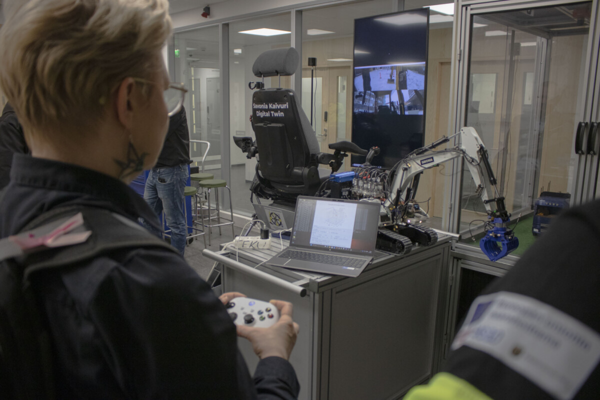 Lyhythiuksinen henkilö pitelee peliohjainta ja käyttää kannettavaan tietokoneeseen liitettyä robottikättä. Laitteen takana on suuri näyttö, joka näyttää kameranäkymän. Huone vaikuttaa modernilta laboratoriolta tai työtilalta.