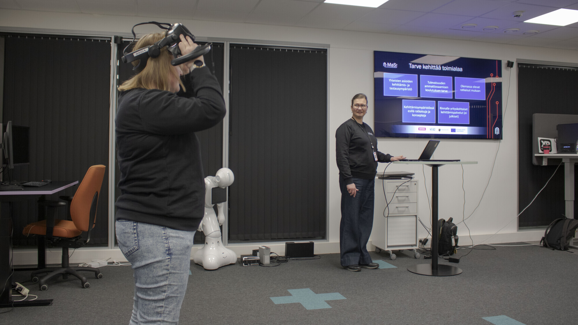 Nainen käyttää virtuaalitodellisuuskuulokkeita, kun toinen henkilö seisoo suuren näytön vieressä pitämässä esitelmää modernissa toimistossa, jossa on pöytiä, tietokoneita ja taustalla robotti.