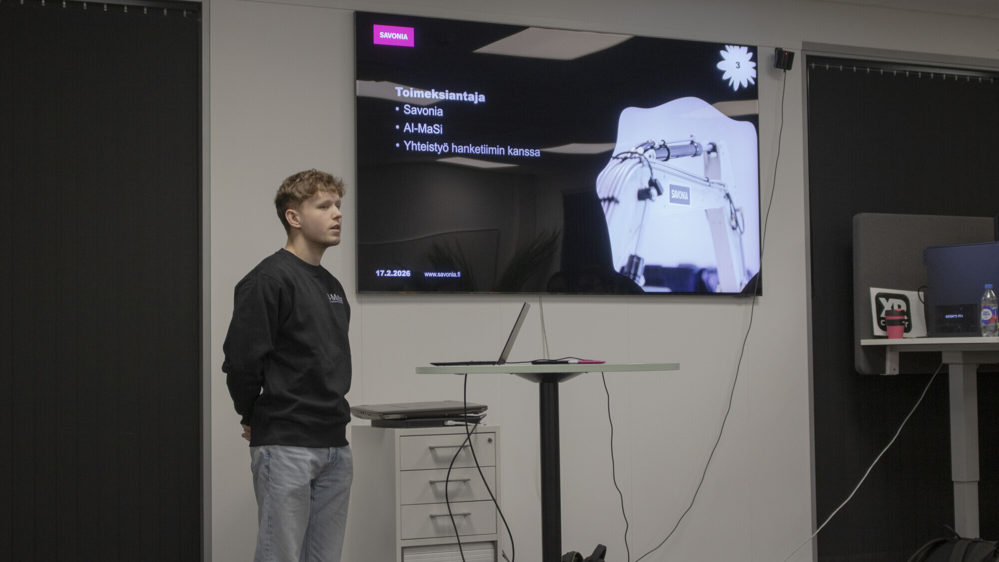 Nuori mies seisoo suuren näytön vieressä, jossa näkyy suomenkielinen esitys. Näytöllä näkyy tekstiä ja kuva robottikädestä, ja mies näyttää puhuvan yleisölle.