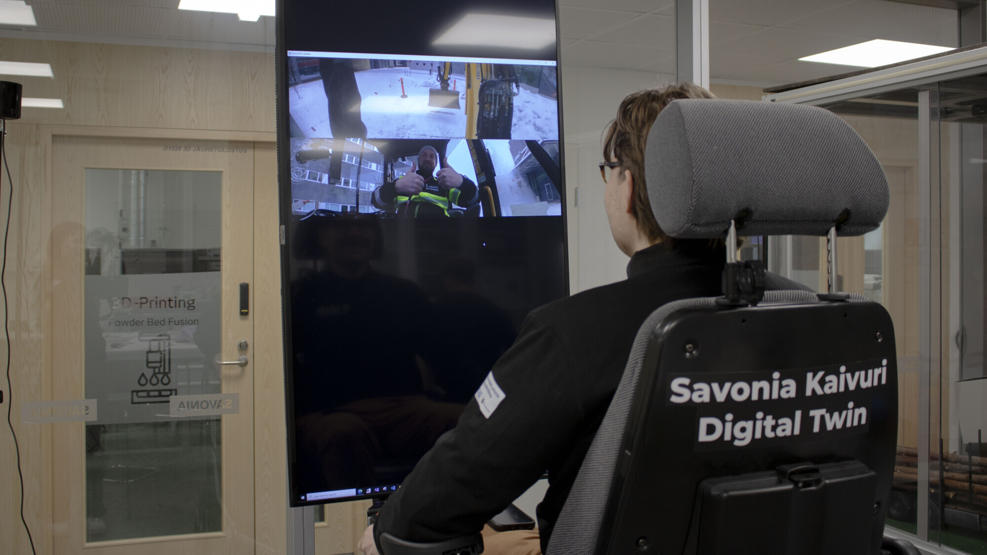 Henkilö istuu ohjaustuolissa, jonka edessä on suuri pystysuora näyttö, jossa näkyy useita kameroita. Paidan selässä lukee Savonia Kaivuri Digital Twin. Ympäristö on moderni toimisto, jossa on lasiseinät ja -ovet.