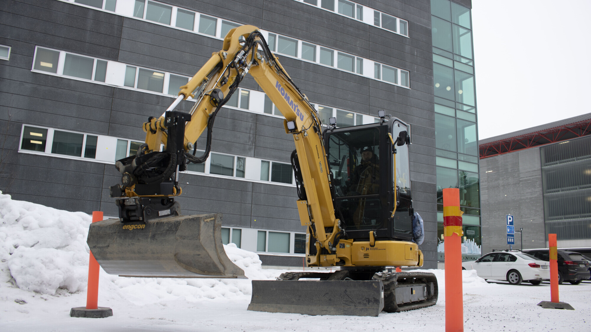 Keltainen kaivinkone työskentelee lumisella rakennustyömaalla modernin harmaan rakennuksen vieressä. Kaivinkoneen varsi on ojennettu, ja sen ympärillä on oransseja turvapylväitä. Taustalla näkyy lunta ja pysäköityjä autoja.