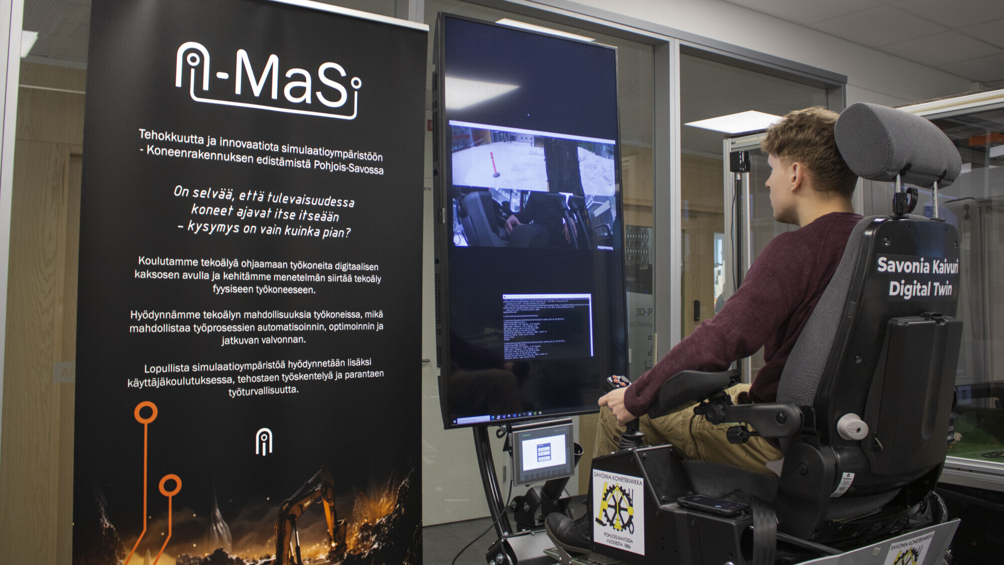 Nuori mies istuu simulaattorituolissa ja katsoo suurta pystysuoraa näyttöä, jossa on teknisiä tietoja. Hänen vasemmalla puolellaan on musta banneri, jossa on A-MaS-nimisen hankkeen logo ja teksti. Ympäristö näyttää olevan moderni laboratorio tai tekninen tila.