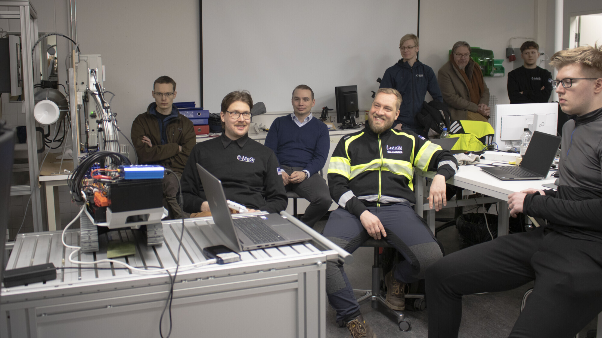 Joukko ihmisiä istuu ja seisoo laboratoriossa tai luokkahuoneessa ja katselee jotain kannettavalta tietokoneelta. Huoneessa on näkyvissä erilaisia teknisiä laitteita ja tietokoneita. Kaikki näyttävät sitoutuneilta ja kiinnostuneilta.
