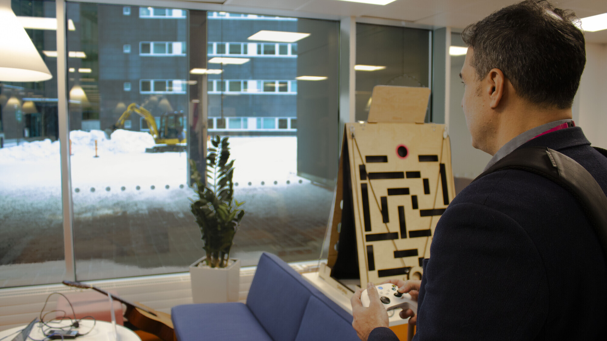 Mies seisoo sisätiloissa peliohjain kädessään ja katsoo suureen ikkunaan. Ulkona lumisella maalla näkyy rakennusajoneuvo. Sisällä lähellä on kasvi, sininen sohva ja puinen labyrinttipeli.