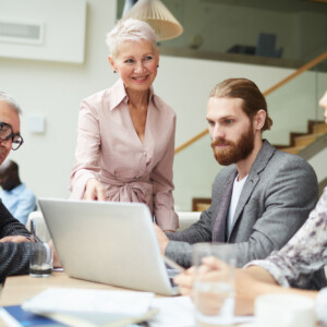 Neljä ammattilaista on kerääntynyt pöydän ääreen kannettavan tietokoneen, asiakirjojen ja vesilasien kanssa. Vanhempi nainen seisoo hymyillen ja osoittaen kannettavaa tietokonetta, kun taas kolme kollegaa istuu ja kuuntelee tarkkaavaisesti.