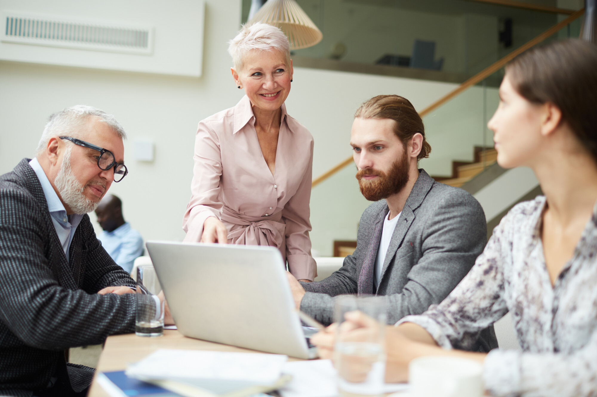 Neljä ammattilaista on kerääntynyt pöydän ääreen kannettavan tietokoneen, asiakirjojen ja vesilasien kanssa. Vanhempi nainen seisoo hymyillen ja osoittaen kannettavaa tietokonetta, kun taas kolme kollegaa istuu ja kuuntelee tarkkaavaisesti.