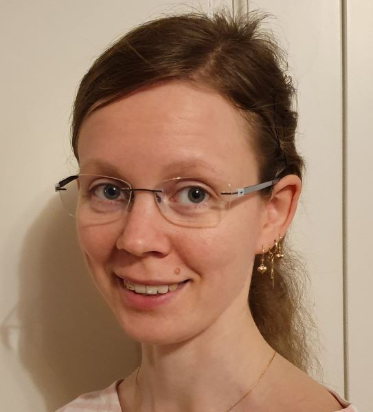 Vaaleaihoinen, silmälasipäinen nainen, jolla on pitkät ruskeat taaksepäin vedetyt hiukset, hymyilee kameralle. Hänellä on kultaiset roikkuvat korvakorut ja vaalea toppi, ja hän seisoo valkoisen seinän edessä.