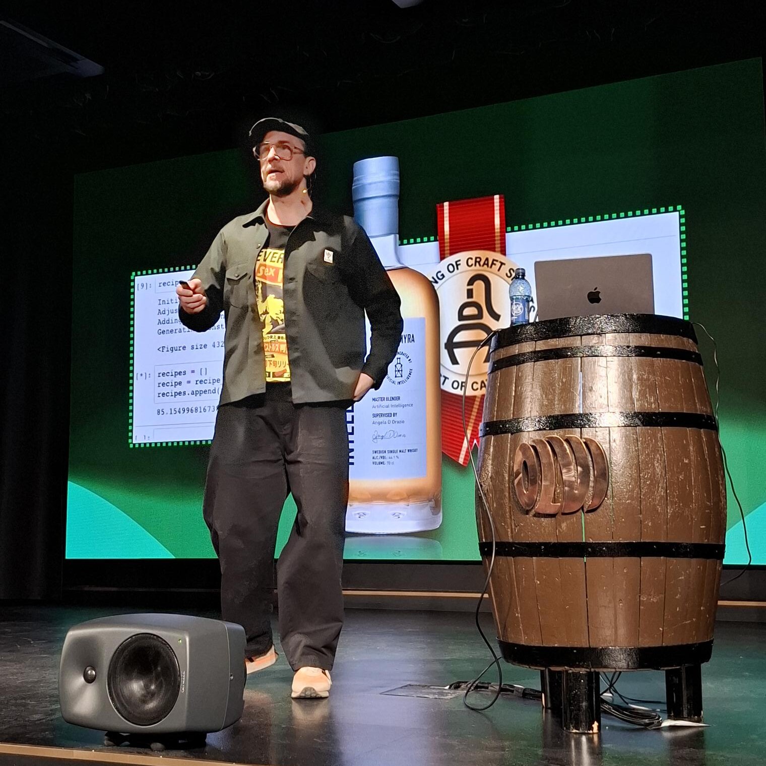 Henkilö puhuu lavalla suuren näytön edessä, jossa näkyy viskipullo. Lattialla on puinen tynnyri, jonka päällä on MacBook ja vesipullo, ja kaiutin. Lavan tausta on vihreä.